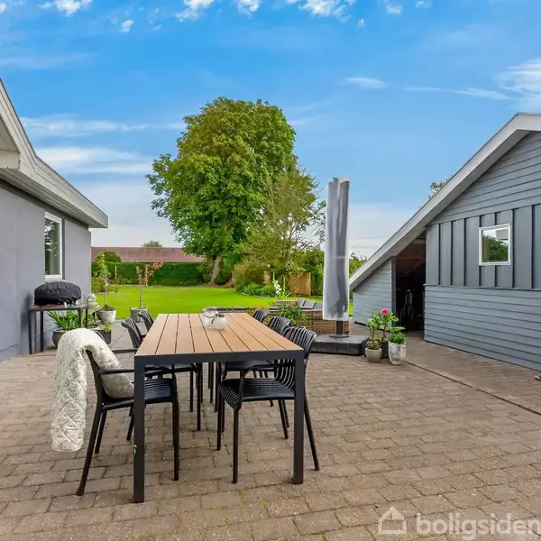 Terrasse med træbord og stole står midt på flisebelagt område. Omkring er et gråt hus, planter og en grill. Baggrunden viser en velplejet have med store træer under en blå himmel.