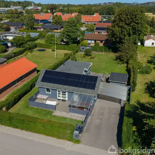 Et hus med solpaneler på taget, omgivet af en grøn have og en indkørsel. Bagskærmen viser kvarteret med flere huse og træer.