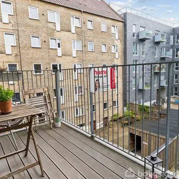 Balkonen har en lille træbord med to stole og en plante. Baggrunden viser et gårdområde omgivet af lejlighedsbygninger. Et skilt fra "home" hænger på en af bygningerne.