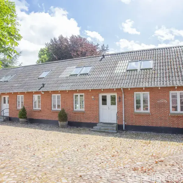 Rødt murstenshus med gråt sadeltag står stille i solrigt vejr. Foran huset er en brostensbelagt gårdsplads med grønne træer på begge sider og krukker med planter.