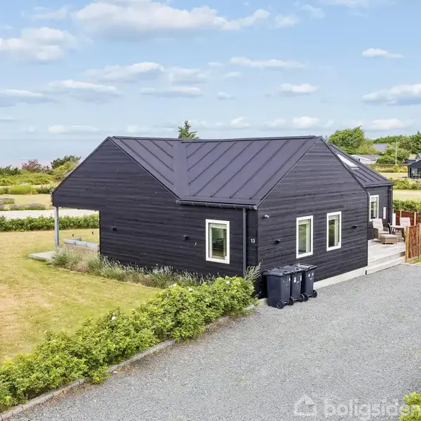 Sort hus med tag i metal, placeret i en grus-indkørsel omgivet af grønne buske. I baggrunden ses flere huse og åbne grønne områder under en skyet himmel.