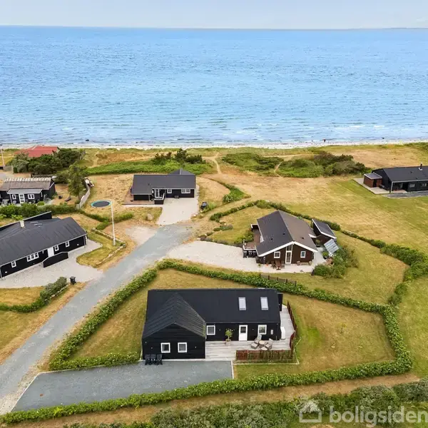 Flere kystnære sommerhuse står stille, omgivet af græsplæner og lave buske; bagved ses stranden og det åbne hav ved horisonten.