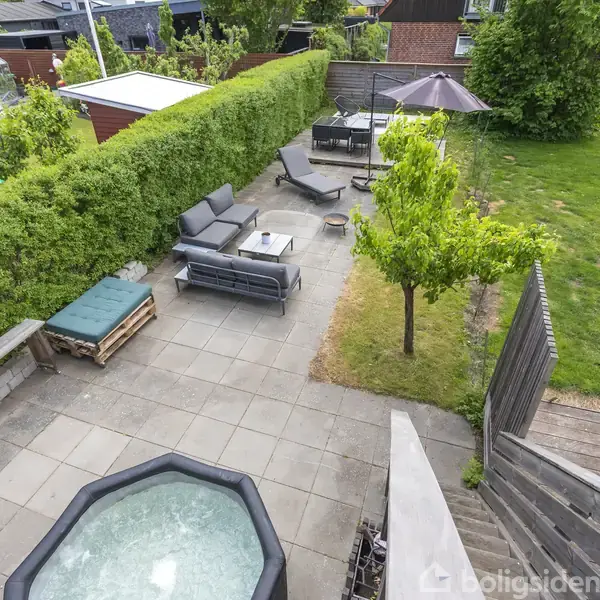 Boblebad står i centrum på en terrasse med fliser; ved siden af er der loungemøbler, træer og en grøn græsplæne. Omkring er der hække og et skur i baggrunden.