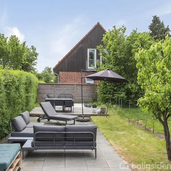 Havemøbler står på en terrasse, flankeret af en grøn hæk og græsplæne. Et stort træhus ses i baggrunden, omgivet af frodig vegetation og trækroner.
