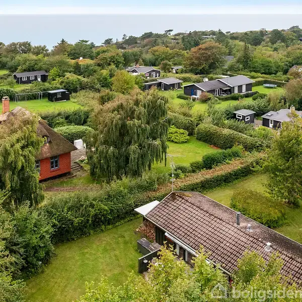 Flere huse ligger spredt i en frodig, grøn havekoloni med tæt vegetation og velholdte græsarealer, omgivet af træer og buske. I baggrunden ses havet og skovklædte områder.