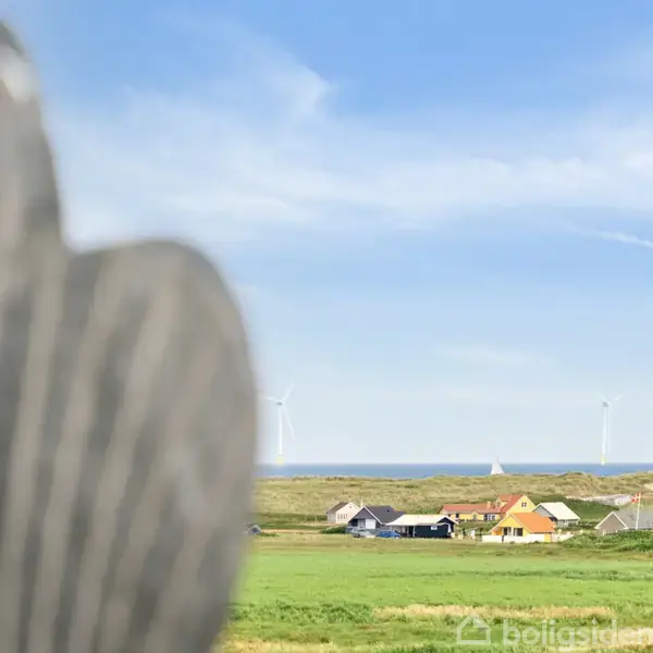 En uklar genstand i forgrunden, sandsynligvis en træskulptur, står foran en landskab med grønne marker, en landsby med farverige huse og vindmøller i baggrunden under en blå himmel.