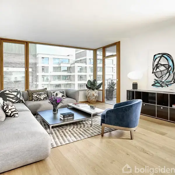 Lys stue med grå hjørnesofa, sofabord og blå lænestol. Store vinduer bagved giver udsigt til moderne bygninger. Et sort opholdsmøbel med dekorationer står på højre side.