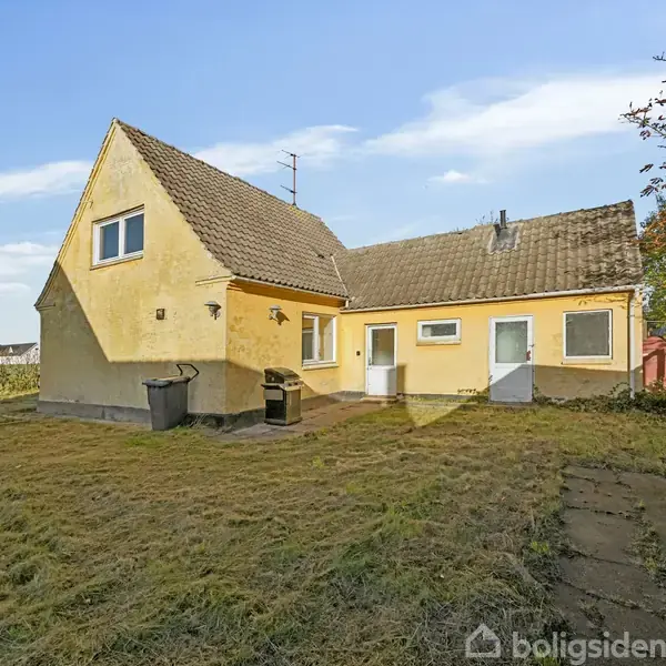 Et gult hus med skråtag står roligt i en have med klippet græs. I baggrunden ses træer. Der er to affaldsbeholdere ved husets side.