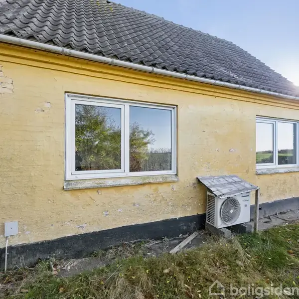 Gul murstensbygning med to vinduer, udluftningsanlæg på ydersiden. Græs voksende omkring, andre huse i baggrunden. Klart vejr, sollys skinner fra højre side.