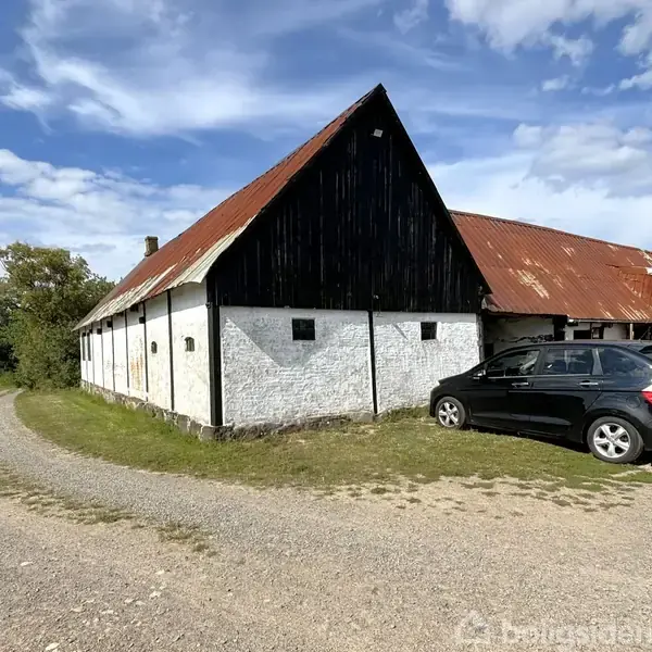 En sort bil holder parkeret ved en gammel hvid bondegård med rødt tag. En grusvej leder op til bygningen under en blå himmel med skyer. En hund står tæt på bilen.