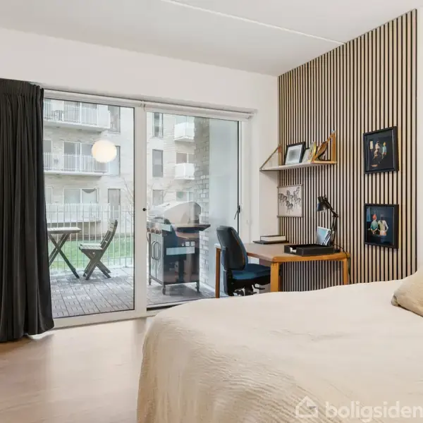 Soveværelse med en seng i forgrunden, et skrivebord langs en væg med træpaneler, og en altan med stole udenfor store vinduer.