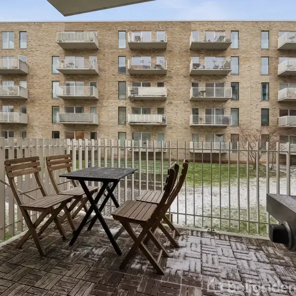 Træbord og stole står på en terrasse, omkranset af et lavt hegn. I baggrunden ses en murstensbygning med flere altaner. Terrassen har også en grill i højre hjørne.