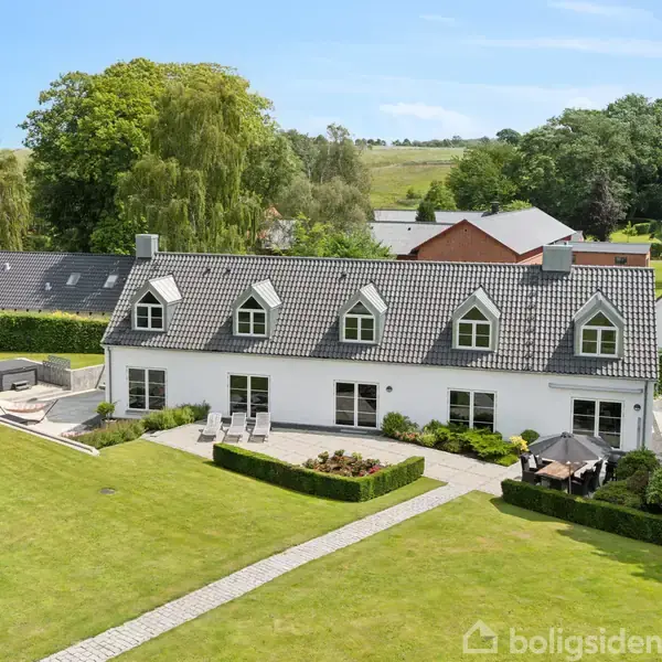 Et stort, hvidt hus med gråt tag står i en velholdt have med buske og en flisebelagt terrasse. Omgivet af frodig natur og skov i baggrunden.