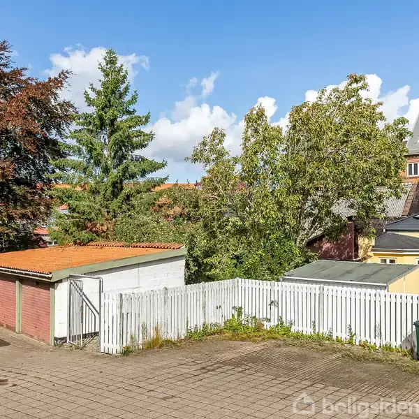 En carport med rød dør står ved siden af et hvidt plankeværk; omgivet af træer og en gul bygning i et boligområde.