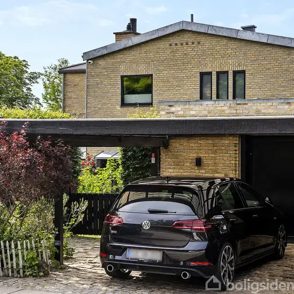 En sort bil holder parkeret under en overdækket carport ved en moderne, gulstensvilla. I baggrunden ses grønne buske og et lyst trægærde.