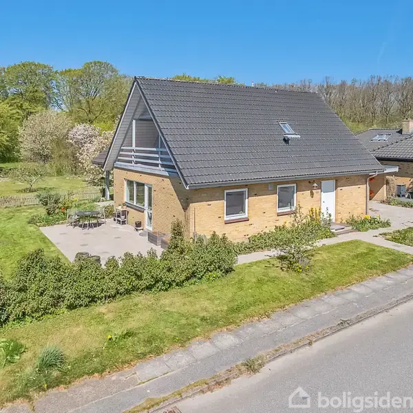 Huset er et gulstenshus med sort sadeltag, omgivet af en velholdt have og terrasse. Det er beliggende i et boligområde med flere træer i baggrunden.