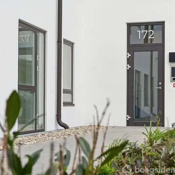 Dør med tallet "172" står i en moderne bygning. Terrassen er omkranset af grus og planter, og der ses flere vinduer langs den hvide facade.