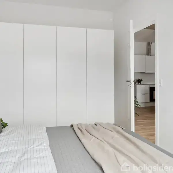 Et soveværelse med en seng dækket af grå og beige sengetøj. En åben dør fører til et køkken. En hvid garderobe langs væggen og en simpel kunstplakat hænger på væggen.