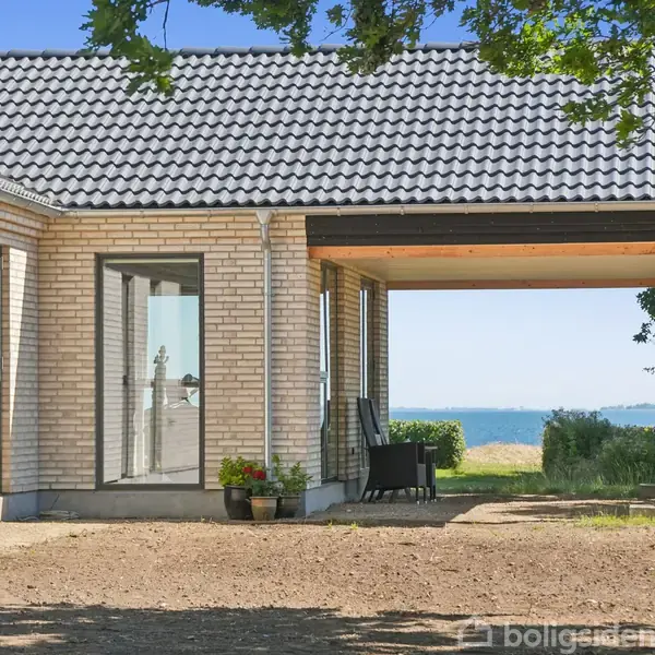 Hus med lyse mursten og grå tagsten. Der er glasdøre, som åbner ud til en overdækket terrasse med havudsigt. Der er græsplæne og træer omkring.