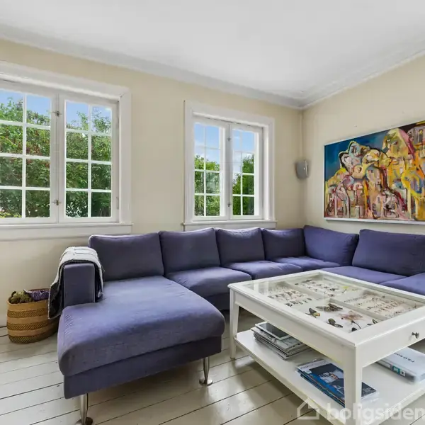 En moderne stue med en blå hjørnesofa placeret mod lyse vægge. Sofabordet i glas og hvid står foran sofaen. På væggen hænger et farverigt maleri. To vinduer med grøn udsigt.