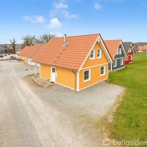 Et gult hus med rødt tegltag står langs en grusvej, omgivet af grønne græsarealer. Andre huse med lignende stil kan ses i baggrunden under en blå himmel.