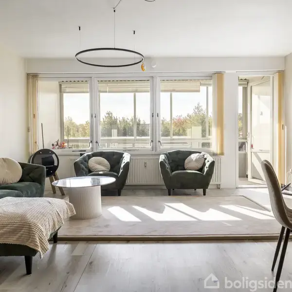 Stue med grøn sofa og lænestole omkring et rundt sofabord. Store vinduer tillader lys at strømme ind, og der er en dør til en balkon i baggrunden. En fladskærms-tv står til højre.