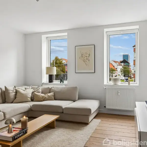 Beige hjørnesofa står i en lys stue med trægulve. Et lavt sofabord bærer pynt. To store vinduer med udsigt til bygninger lukker lys ind.