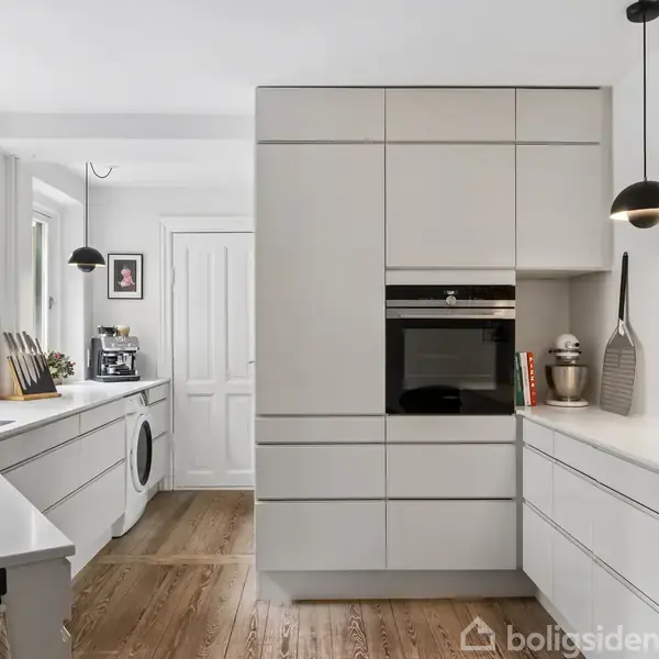 Moderne køkken med hvide skabe og indbygget ovn. Belysning fra hængelamper, stilfulde bordplader og en vaskemaskine i baggrunden. Naturligt lys kommer ind fra vinduet.