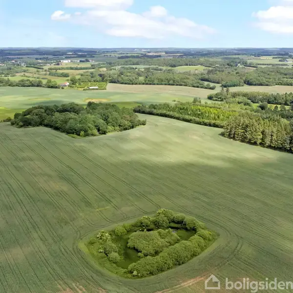 Grønne marker strækker sig over et bølgende landskab med enkelte skovområder. Himlen er klar med spredte skyer, hvilket skaber en rolig atmosfære.