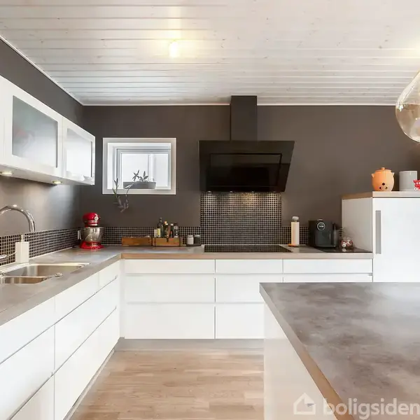Et moderne køkken med hvide skabe og stålplader, hvor en elektrisk ovn og en emhætte er installeret. En stor, pæreformet lampe hænger over bordet.
