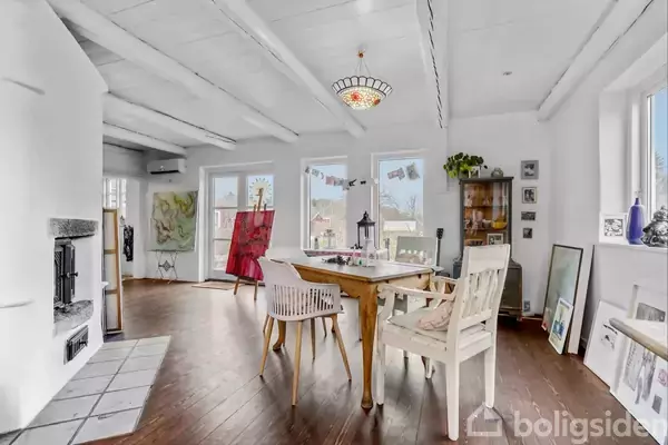 Et lyst rum med hvide vægge og træloft. Et træbord og stole står midt i rummet, omgivet af malerier og planter ved vinduerne. Ingen tekst fundet.