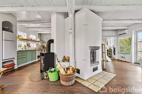 Brændeovn i rum med hvidmalede vægge, står midt i et åbent køkken-alrum. Rummet har trægulv, kurve med brænde, grønne køkkenelementer, og lysindfald fra flere vinduer og en glasdør.