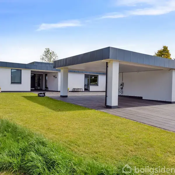 Et moderne, hvidt hus med fladt tag står stille ved en velplejet græsplæne og flisebelagt terrasse. Baggrunden viser en blå himmel og grønne buske.