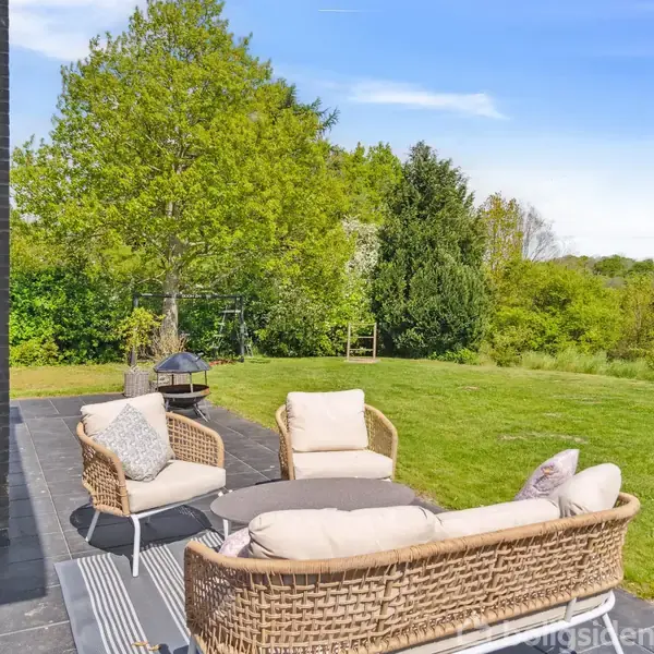 Rattanmøbler står på en terrasse med sorte fliser, omkranset af en grøn græsplæne og træer i baggrunden. Solen skinner klar på en skyfri blå himmel.