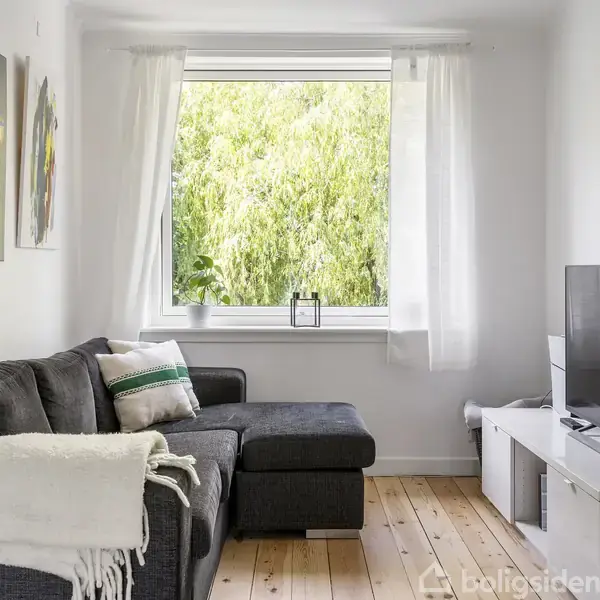 Grå hjørnesofa med tæppe og puder står i en stue med trægulv. Store vinduer med udsigt til grønne træer. Fladskærms-TV på hvidt bord. Vægkunst til venstre.