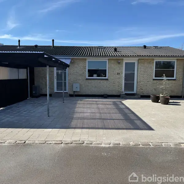 En enkel etplans murstensvilla med en carport til venstre og flisebelagt indkørsel foran. Huset har et vindue og en dør med tre urtepotter til højre.