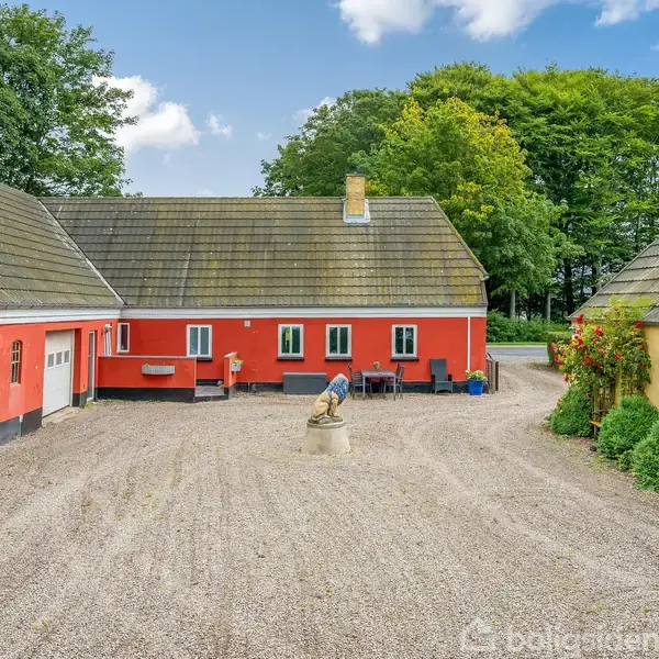 En rød bondegård omgivet af en grusgård med grønne træer i baggrunden. Til venstre står en garagebygning, og til højre et gult udhus med planter langs væggen.