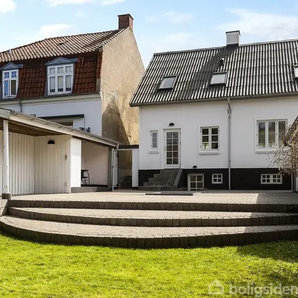 Hvidt hus med trappe fører til en terrasse. Græsplæne og carport til venstre. Nabobygning med rødt tag. Klar blå himmel over.