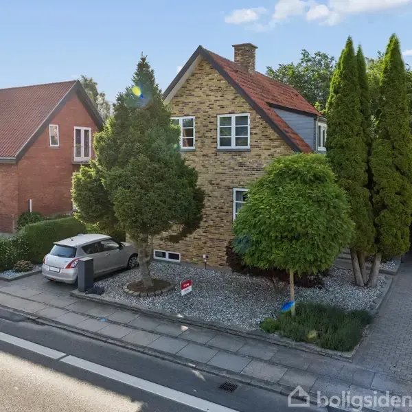 En murstensvilla med rødt tag og hvide vinduer står omgivet af grønne træer. En sølvfarvet bil er parkeret på indkørslen ved siden af huset, som ligger i et villakvarter.