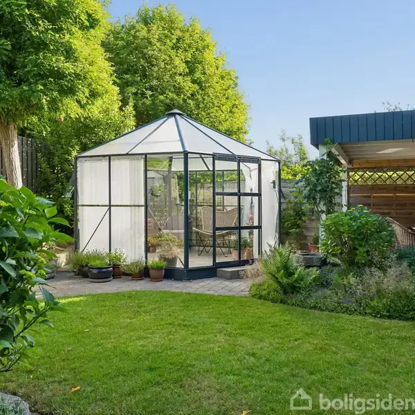 Et fritstående drivhus med glasvægge står i en velplejet have omgivet af grønt løv. Ved siden af ses en åben carport. Græsplænen strækker sig om drivhuset.