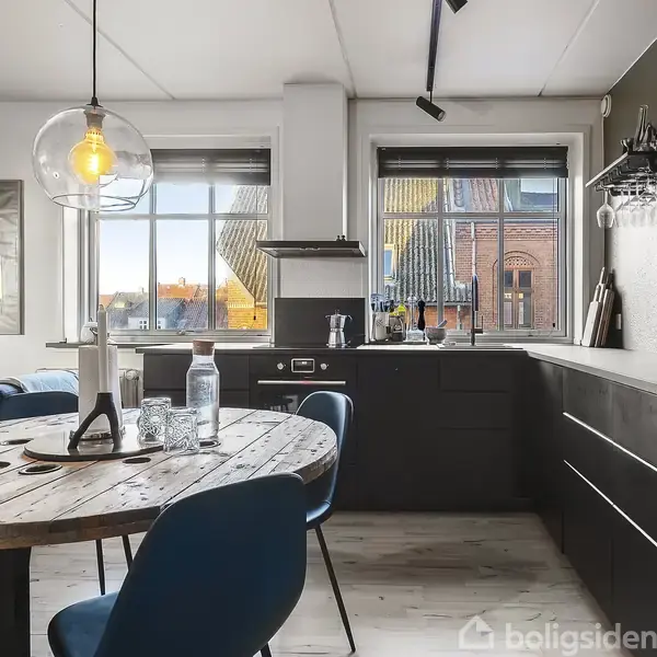 Rundt spisebord med blå stole står i et moderne køkken med sorte skabe. Store vinduer giver udsigt til farverige bygninger udenfor. En dekorativ lampe hænger over bordet.