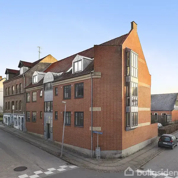 Rød murstensbygning med skråtag og store vinduer står ved et vejkryds. Flere biler er parkeret langs vejen, som er omgivet af andre ældre bygninger i et byområde.
