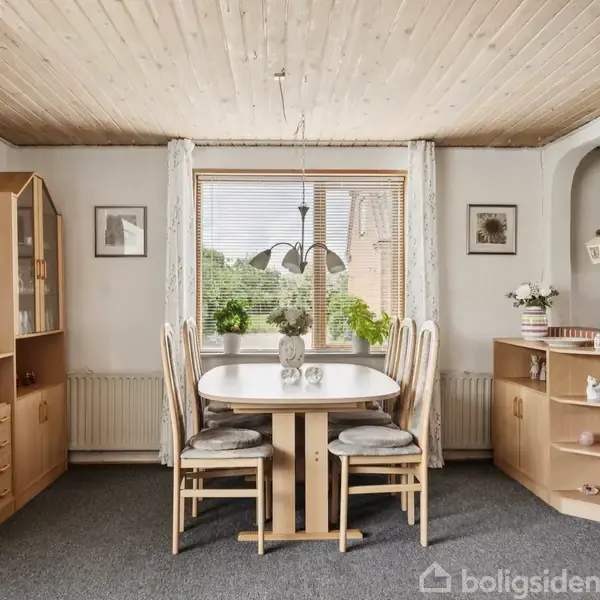 Spisebord med fire stole står i en stue med trægulv og loft. Omgivet af brune reoler og en buet murstensvæg til højre. Store vinduer giver udsigt til grønne omgivelser.