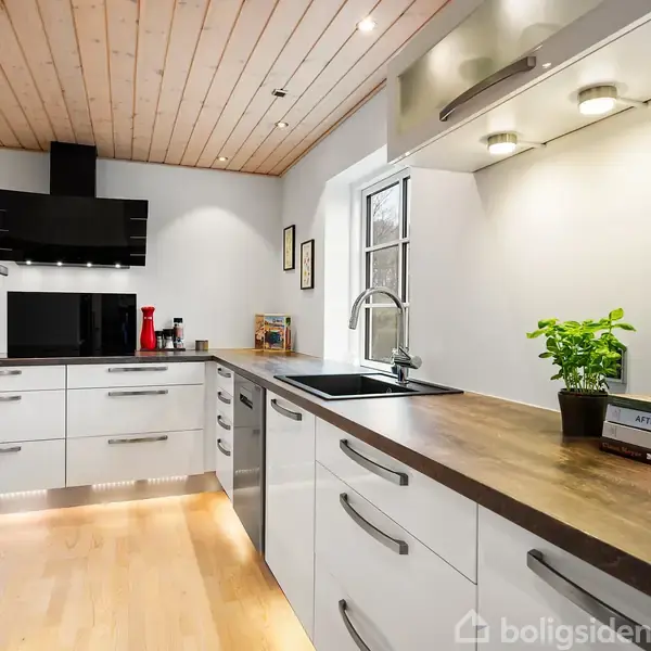 Et moderne køkken med hvide skabe udfører opbevaring, mens en håndvask er integreret i en træbordplade. En grøn plante og bøger er placeret ved siden af vinduet.