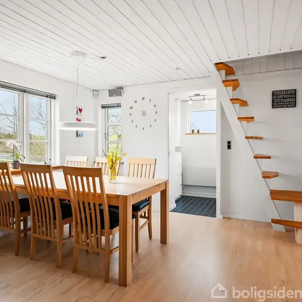 Et spisebord med træstole står i en lys spisestue ved vinduer. En trappe uden gelænder fører opad mod højre. På væggen ses et ur og et skilt med teksten "Livet er et eventyr der skal leves - forsømmelse af det kan medføre savn på sjælen".