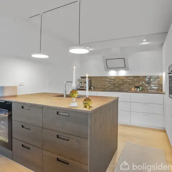 Køkkenø med mørke skuffer står centralt; to pendellamper hænger over. I baggrunden er der hvide skabe og en blank flisevæg. Der er en indbygget ovn og en vinkøler.