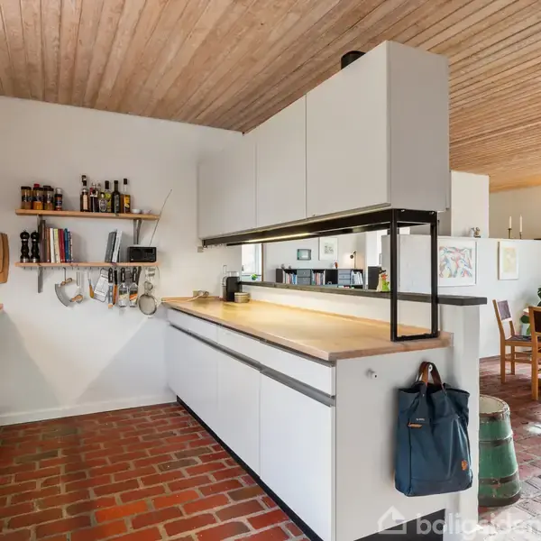 Køkken med hvide skabe og træbordplade, udstyret med kogeplader og vægophængte hylder, ligger i et rum med flisegulv af røde mursten. Køkkenet åbner op til en lys spiseplads.