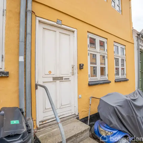 Gul bygning med ældre facade prydet af hvide døre og vinduer. Fortovet har trapper, rækværk og blomsterkasser. Nummer 32 vises over døren. To affaldscontainere står nær indgangen.