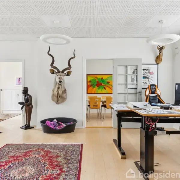 Et kontor med et skrivebord fyldt med papirer. En computer står på bordet. Væggene har kunst og en hjortetrofæ. Et gulvtæppe dækker trægulvet. I baggrunden ses en statue og malerier.