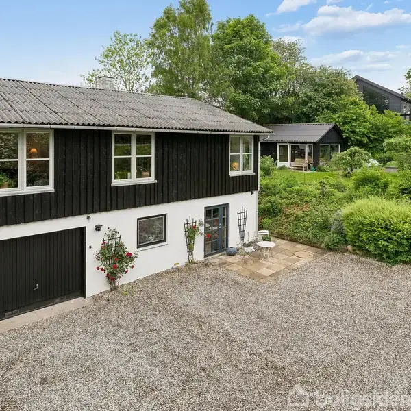 To-etagers hus med sort og hvid facade. Grusindkørsel foran, omkranset af grønne træer og buske i haveområde. Der er en trappe på siden, der fører til øverste etage.